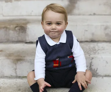 A young child in a navy sweater vest with a festive design, sitting on stone steps, smiling.