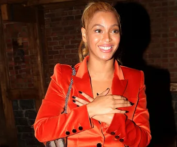Woman smiling in a red blazer with hands crossed on chest, standing in front of a brick wall.
