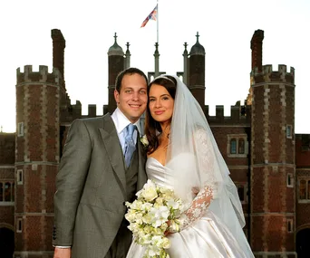 "Couple in wedding attire posing in front of a historic building with a flag flying above."