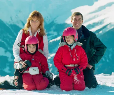 A family of four in winter clothing poses on a snowy mountain with a clear blue sky background.