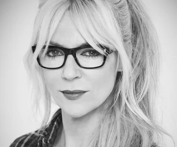 A woman with long blonde hair and glasses, wearing a plaid shirt, in a black and white portrait.
