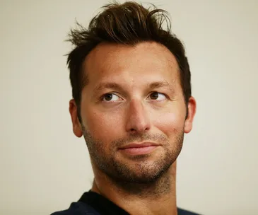 A person with short hair and a slight beard gazes upwards, appearing thoughtful, against a neutral background.