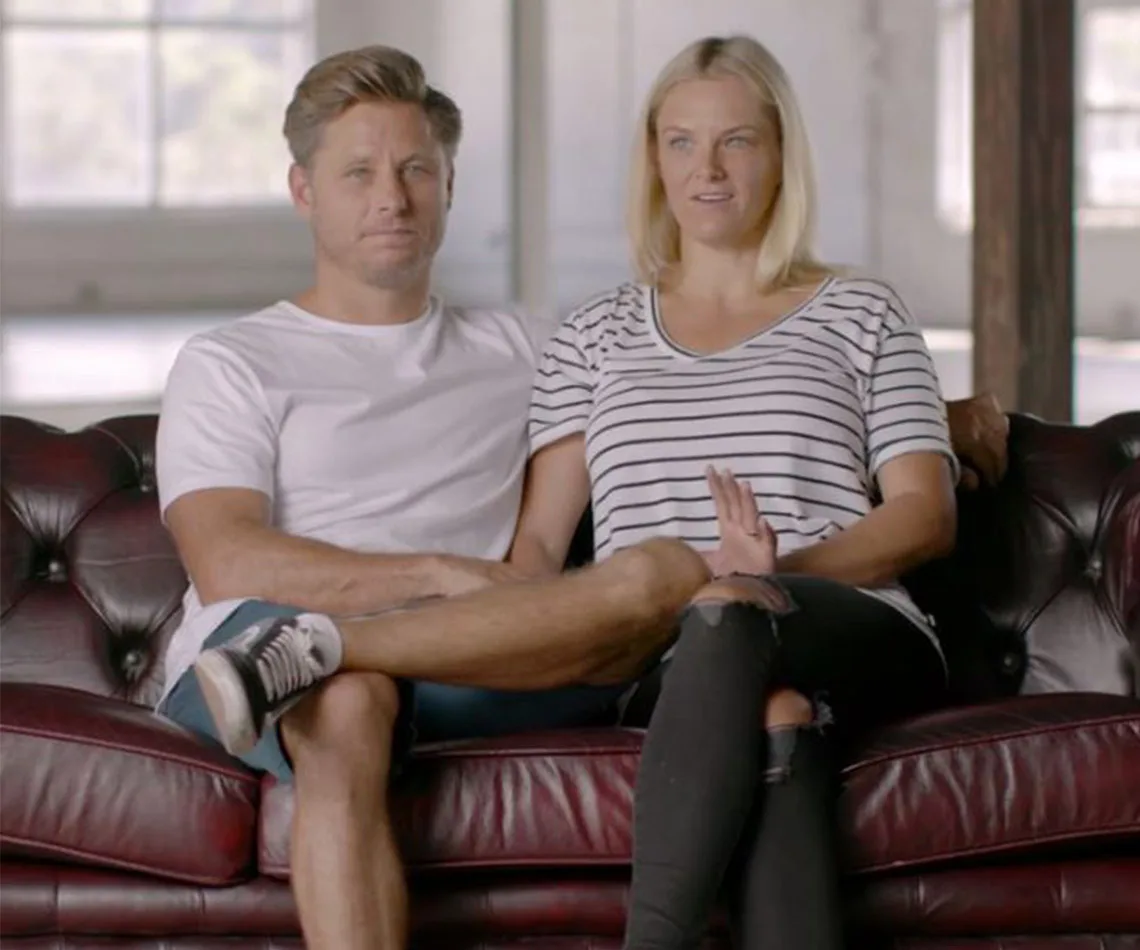 A couple sitting on a red leather couch, smiling and relaxed, in a bright room with large windows in the background.