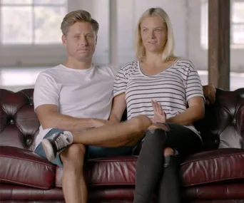A couple sitting on a red leather couch, smiling and relaxed, in a bright room with large windows in the background.