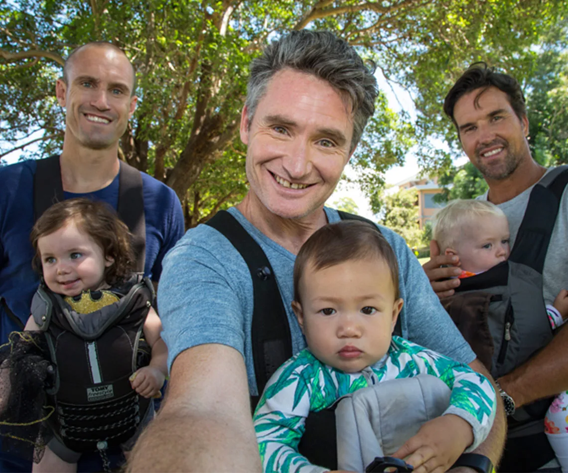 Men holding babies in carriers, smiling in a sunny park setting.