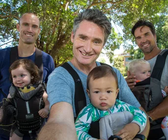 Men holding babies in carriers, smiling in a sunny park setting.