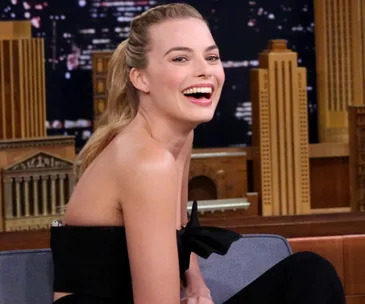 Margot Robbie smiling, sitting on a talk show set, wearing a black dress and showing a wedding ring.