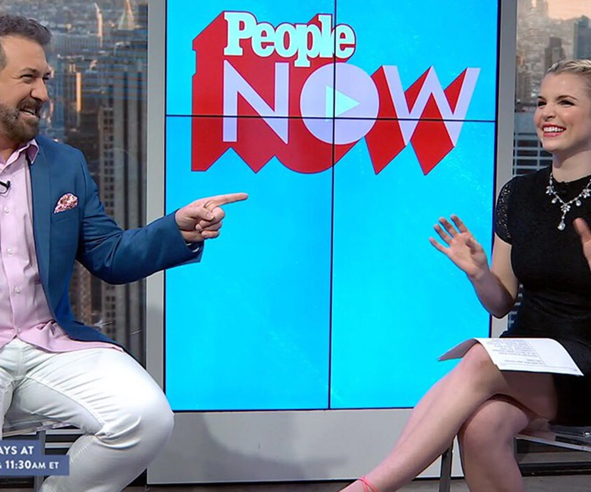 Man and woman talking, seated in front of a "People NOW" screen, in a studio setting with cityscape views.