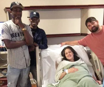 Group in a hospital room: two men stand on the left, a woman in a hospital bed smiles, and a man on the right touches her arm.