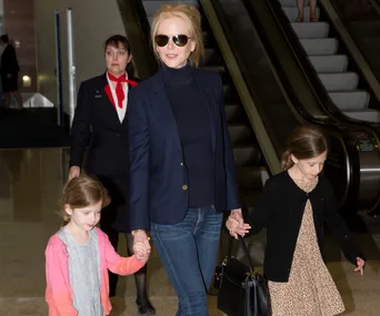 Nicole Kidman, in sunglasses, holding hands with two young girls at an airport, with escalators in the background.