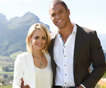 A couple smiling and posing outdoors with mountains in the background, both wearing formal attire.