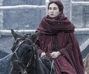 Melisandre from Game of Thrones rides a horse in a snowy setting, wearing a red cloak.