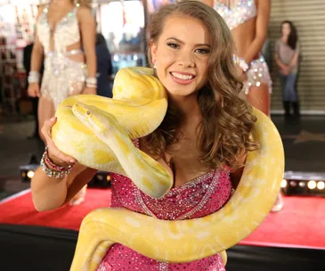 I don't know who this is, but she's holding a large yellow snake while smiling, wearing a sparkling pink dress.