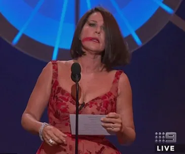 Woman in red dress with smeared lipstick speaking at an event, holding a paper.
