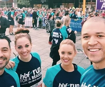 Group of four people smiling at a charity run event, wearing "HBF Run for a Reason" shirts, with a crowd in the background.