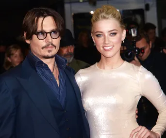 A man in a dark suit and glasses stands beside a woman in a sparkling dress at a public event, with photographers in the background.
