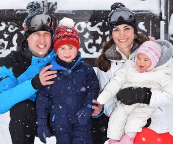 Family in winter attire smiling in the snow, with a young boy and girl wearing colorful jackets and hats.
