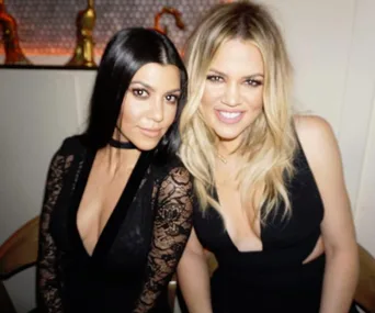 Two women in black dresses smiling at the camera, seated in a stylish indoor setting.