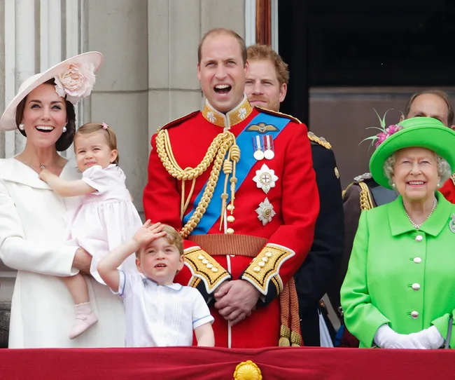A royal family on a balcony, smiling. A child in front salutes while another is held in a woman's arms.