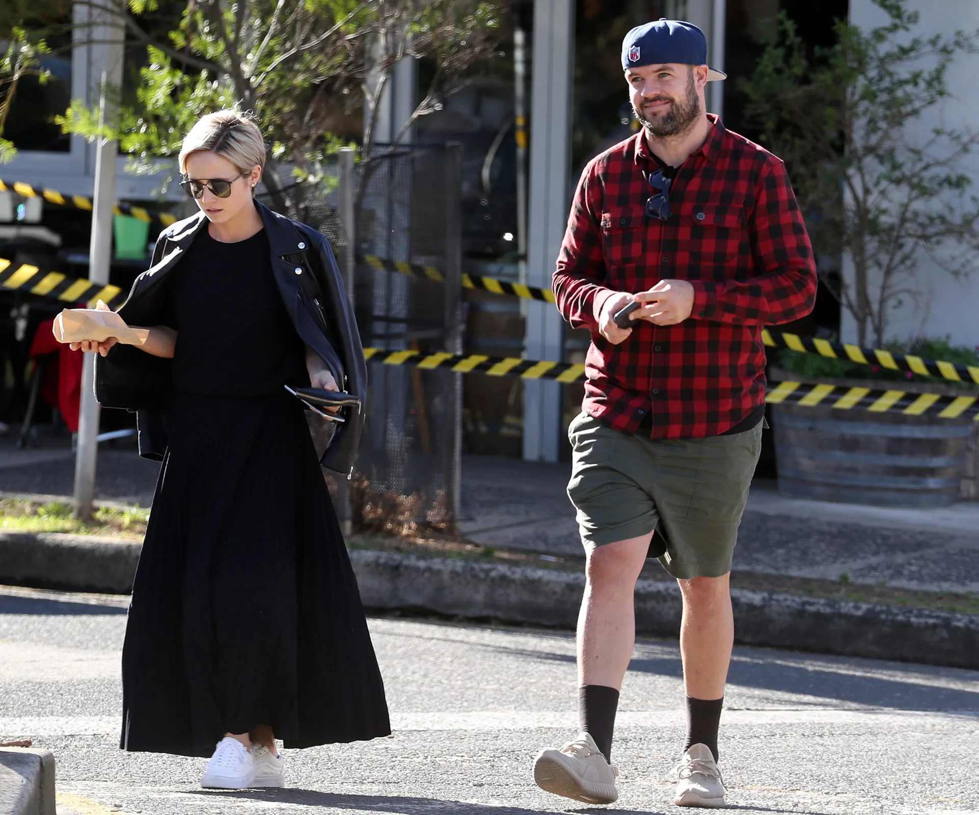Two people walking outdoors, one in a black outfit with sunglasses, the other in a red plaid shirt and cap.