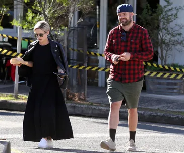 Two people walking outdoors, one in a black outfit with sunglasses, the other in a red plaid shirt and cap.