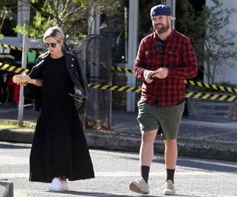 Two people walking outdoors, one in a black outfit with sunglasses, the other in a red plaid shirt and cap.