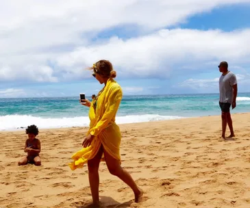 A woman in a yellow cover-up on a beach, child playing in sand, and man looking at the ocean under a bright sky.
