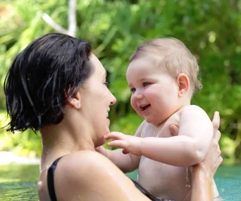 A woman holds a smiling baby in a pool, surrounded by greenery.
