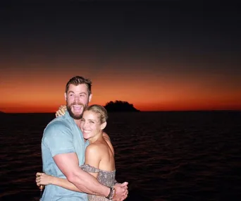A couple embraces joyfully in front of a stunning sunset over the ocean.