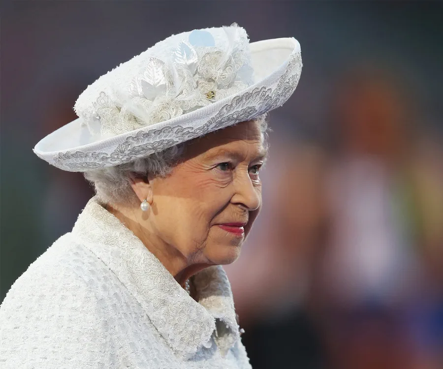 A woman wearing a detailed white hat and outfit with lace and embroidery, featuring pearl earrings.