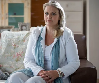 A woman sitting on a couch in a living room, wearing a white outfit and blue scarf.