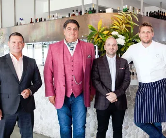 Four men in formal clothes and a chef outfit standing in front of a bar with plants and bottles in the background.