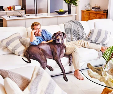 Man lounging on a white sofa with a large gray dog in a modern living room, featuring a kitchen in the background.
