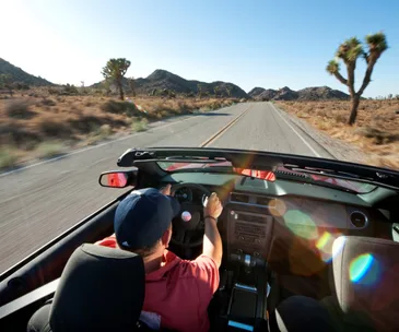 Person driving a convertible car on a scenic desert road in California under a clear blue sky.