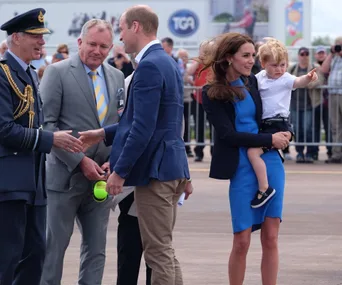 Family at airshow; man in uniform shakes hands, woman holds child pointing, surrounded by people.