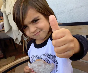 A child with long hair gives a thumbs-up while holding a snack, sitting at an outdoor table.
