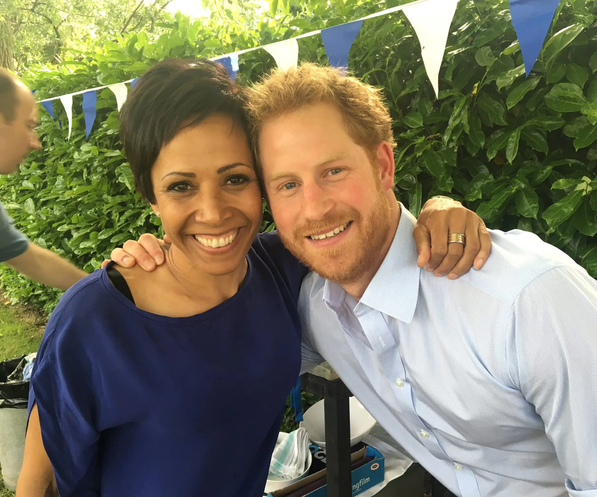Two people smiling at an outdoor event, with blue-and-white bunting and greenery in the background.