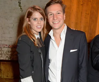Couple smiling at an event, dressed in formal black and white attire, against a brick and wood backdrop.