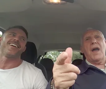 Father and son smiling and singing together in a car during Carpool Karaoke segment.