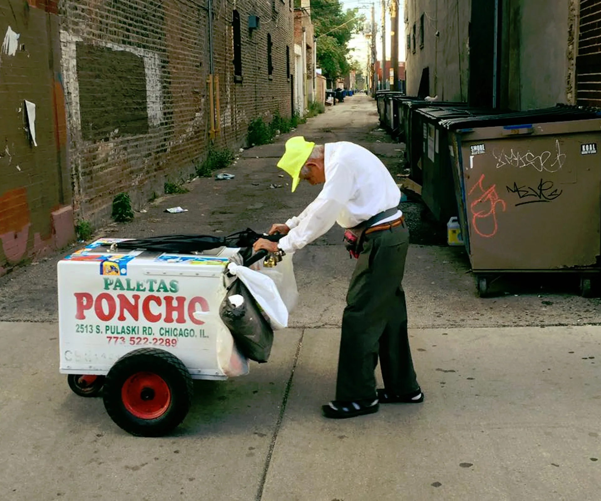 Elderly man pushing an ice cream cart labeled "Paletas Poncho" in an alleyway, wearing a bright yellow cap.