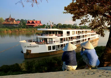 River cruise ship "La Marguerite" passing by traditional buildings, two people in conical hats observing from the shore.