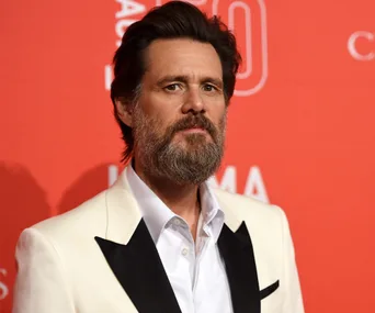 A man with a beard wearing a white suit with a black lapel stands against a red background, looking forward.