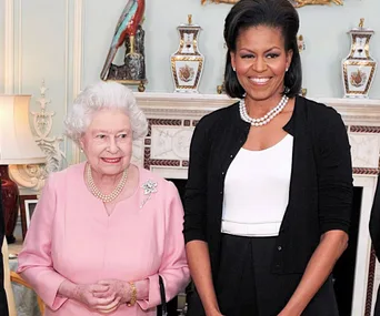 Two women smiling, one in a pink suit and pearls, the other in a black cardigan and white top, standing indoors.