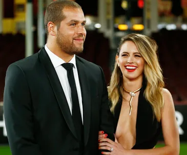 A couple in formal attire, the man in a black suit and tie, the woman in a black dress with gold jewelry, smiling together.