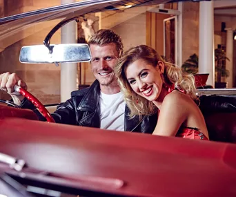 A smiling couple sitting in the front seats of a red vintage car, with the woman showing blonde hair.