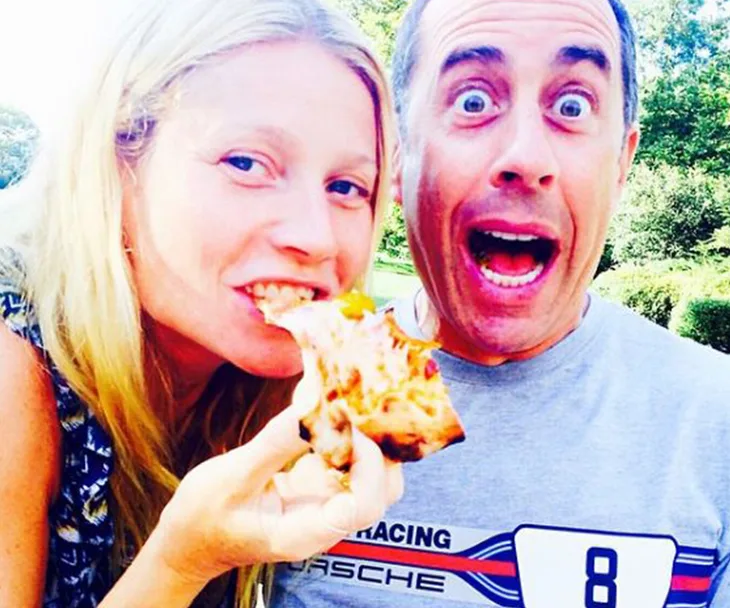 A woman eats pizza while a surprised man looks on, both outdoors and smiling.