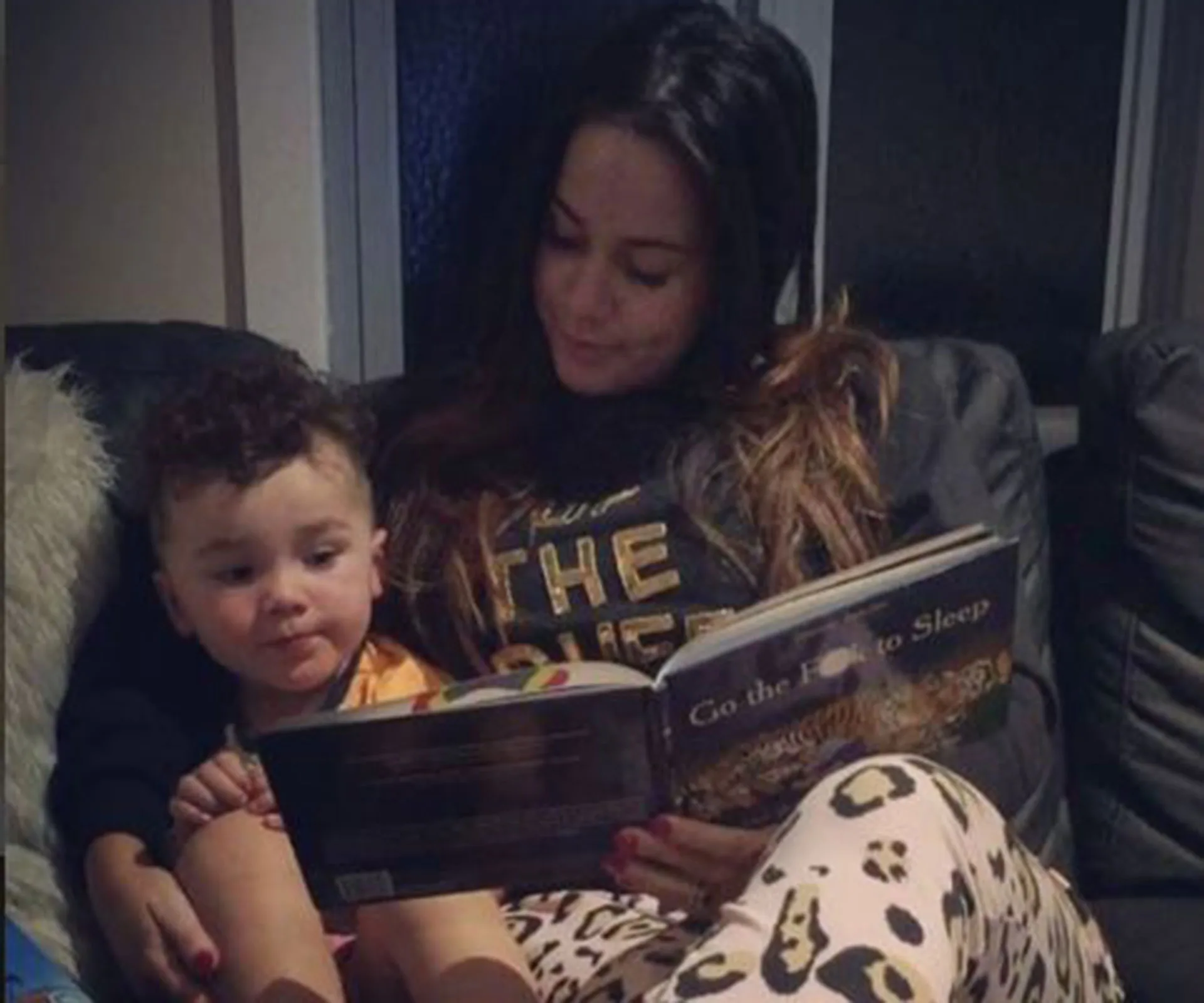 A mother reads "Go the F*** to Sleep" to her child on the couch, both looking at the book intently.
