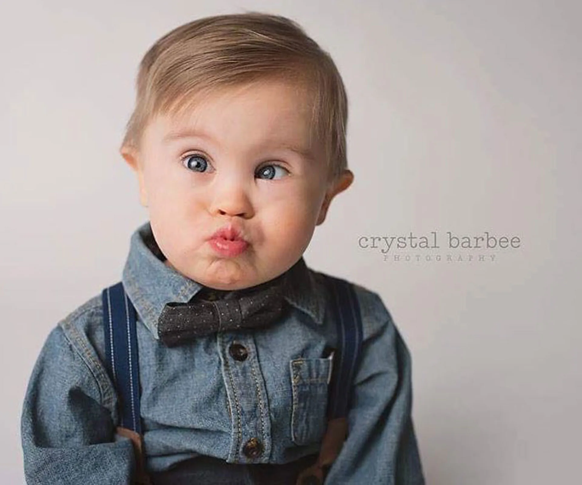 A toddler with Down syndrome wearing a denim shirt and bow tie makes a playful pouty face.