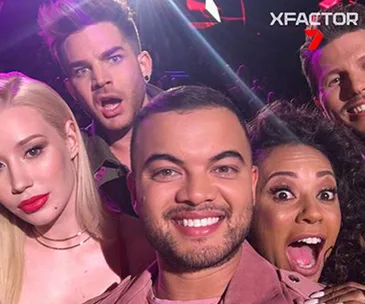 A group of five people taking a selfie, smiling and making excited expressions, with "X Factor" logo in the background.