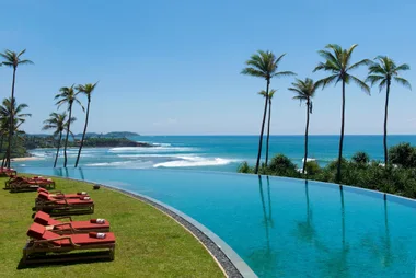 Infinity pool overlooking the ocean with palm trees and lounge chairs on a sunny day in Sri Lanka.